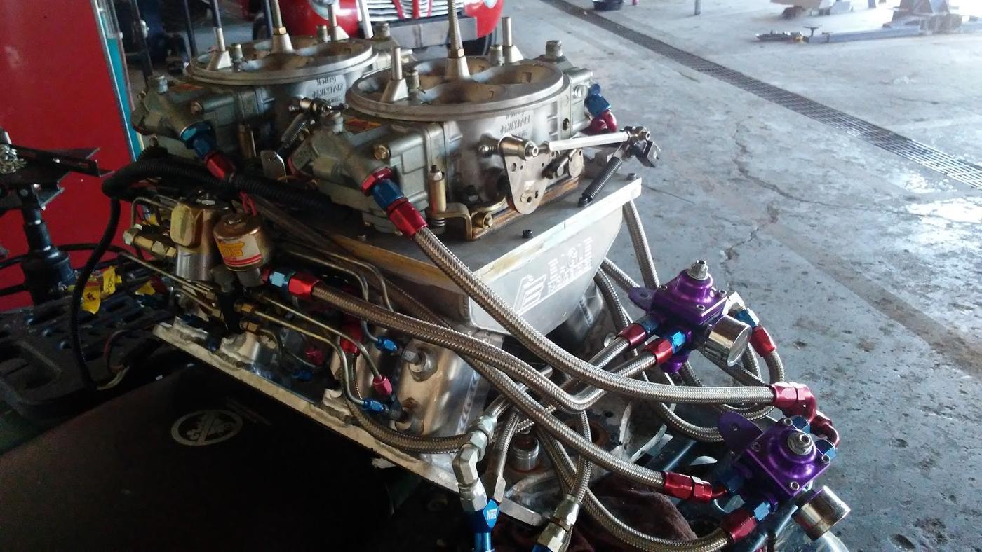 Twin carburetors on a tunnel ram intake, braided fuel lines and regulators, photographed in the shop